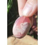Gros plan sur pendentif rhodochrosite ovale rose marbré avec monture argent 925, tenu entre les doigts devant fond végétal et champignon, bijou en pierre naturelle associé à la sensibilité apaisée, à la douceur et à l’équilibre du cœur.