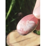 Gros plan sur pendentif rhodochrosite ovale rose marbré serti argenté, tenu entre les doigts avec fond végétal et bois, bijou en pierre naturelle lié au réconfort, à la douceur du cœur et à la stabilité intérieure.