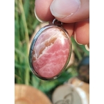 Pendentif rhodochrosite ovale rose marbré en argent 925 tenu entre les doigts devant un fond végétal, pierre naturelle associée à la délicatesse, au recentrage et à la paix émotionnelle.