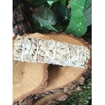Bâton de sauge blanche à brûler (fumigation/smudge) ficelé, posé sur rondin de bois avec feuillage vert et décor naturel. Accessoire pour rituel, méditation, purification énergétique, nettoyage des lieux, protection, clarté et apaisement.