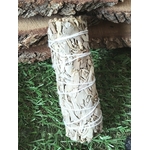 Bâton de sauge blanche à brûler (fumigation/smudge) ficelé, présenté debout sur feuillage vert avec décor de bois. Accessoire pour rituel, méditation, purification énergétique, nettoyage des lieux, protection, clarté et apaisement.