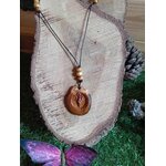 Pendentif bois Gaïa rond gravé suspendu devant tranche de bois avec mousse verte, cordon noir et perles bois dans une ambiance forestière