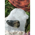 Pierre roulée de serpentine vert olive sur amas de quartz blanc, feuillage en fond; apaisement, ancrage, équilibre émotionnel.