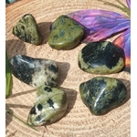 Pierres roulées de serpentine vert olive sur tranche de bois clair, avec plume violette; pierre associée à l’apaisement, à l’ancrage et à l’équilibre émotionnel.