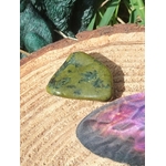 Pierre roulée de serpentine vert olive, sur tranche de bois avec plume violette et feuillage, lumière douce; associée à l’apaisement, à l’ancrage et à l’équilibre émotionnel.