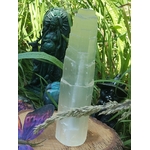 Tour en sélénite blanche posée dans l’herbe, fond nature, corde naturelle devant, plume multicolore et petit élément bleu à côté, sensation de purification douce.