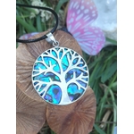 Pendentif arbre de vie en abalone et argent 925 sur cordon noir, posé sur rondin de bois avec herbes vertes et papillon rose flou, bijou en nacre naturelle évoquant la confiance douce, la paix intérieure et la sensibilité apaisée.