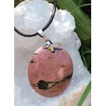 Pendentif rond en rhodonite sur cordon noir, posé sur amas de quartz blanc et feuilles vertes, bijou en pierre naturelle relié à l’apaisement du cœur, à l’ancrage doux et à la sérénité.