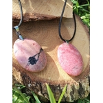 Deux pendentifs en rhodonite rose sur cordon noir, posés sur une rondelle de bois au jardin, bijoux en pierre naturelle associés à la tendresse, à l’équilibre affectif et au calme intérieur.