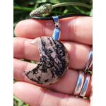 Pendentif lune en rhodonite rose marbrée de noir tenu dans une main, cordon noir et bélière argentée visibles, bijou en pierre naturelle relié à la bienveillance, au pardon et à l’équilibre du cœur.