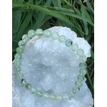 Bracelet en préhnite 6 mm vert d’eau posé sur un amas de quartz blanc, herbes vertes floues en arrière-plan, pour apaiser et clarifier l’esprit.