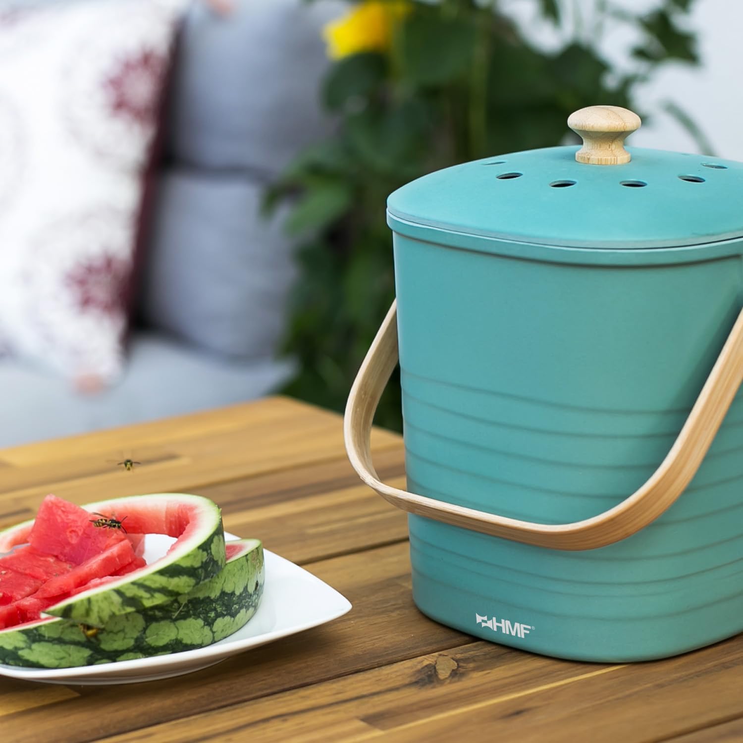 Seau à compost turquoise avec anse en bambou, posé sur une table près dune tranche de pastèque