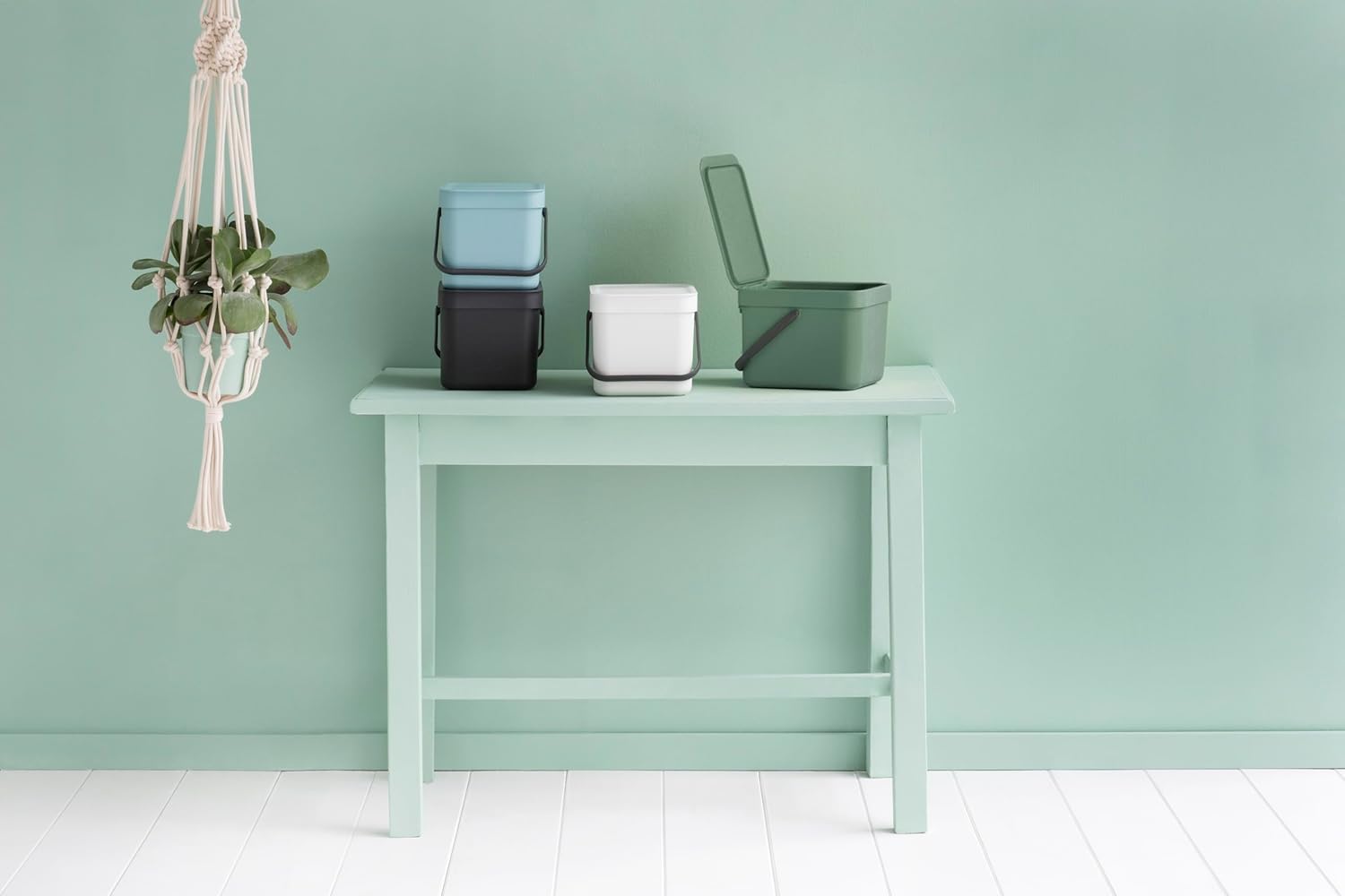 Seaux Brabantia de plusieurs coloris sur une console vert deau, avec une plante suspendue