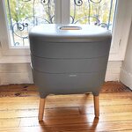 Lombricomposteur de balcon WormBox gris de 35 L, à trois niveaux et pieds en bois, devant une fenêtre