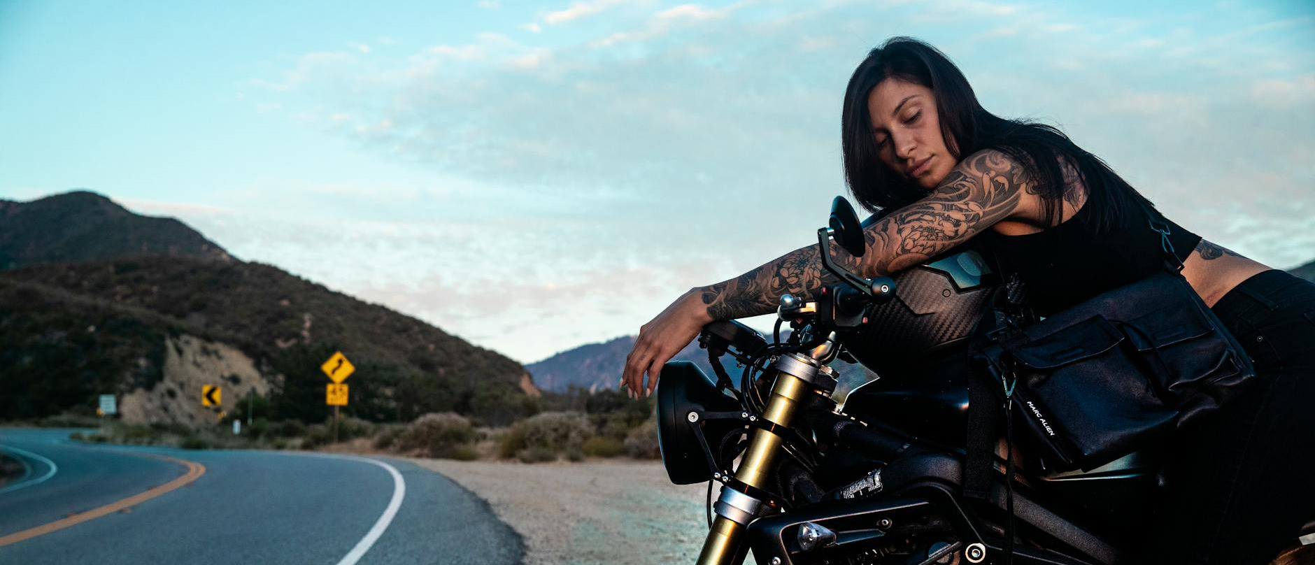 a tattooed woman rests on a motorcycle by a curving road in a tranquil scenic landscape capturing the essence of freedom
