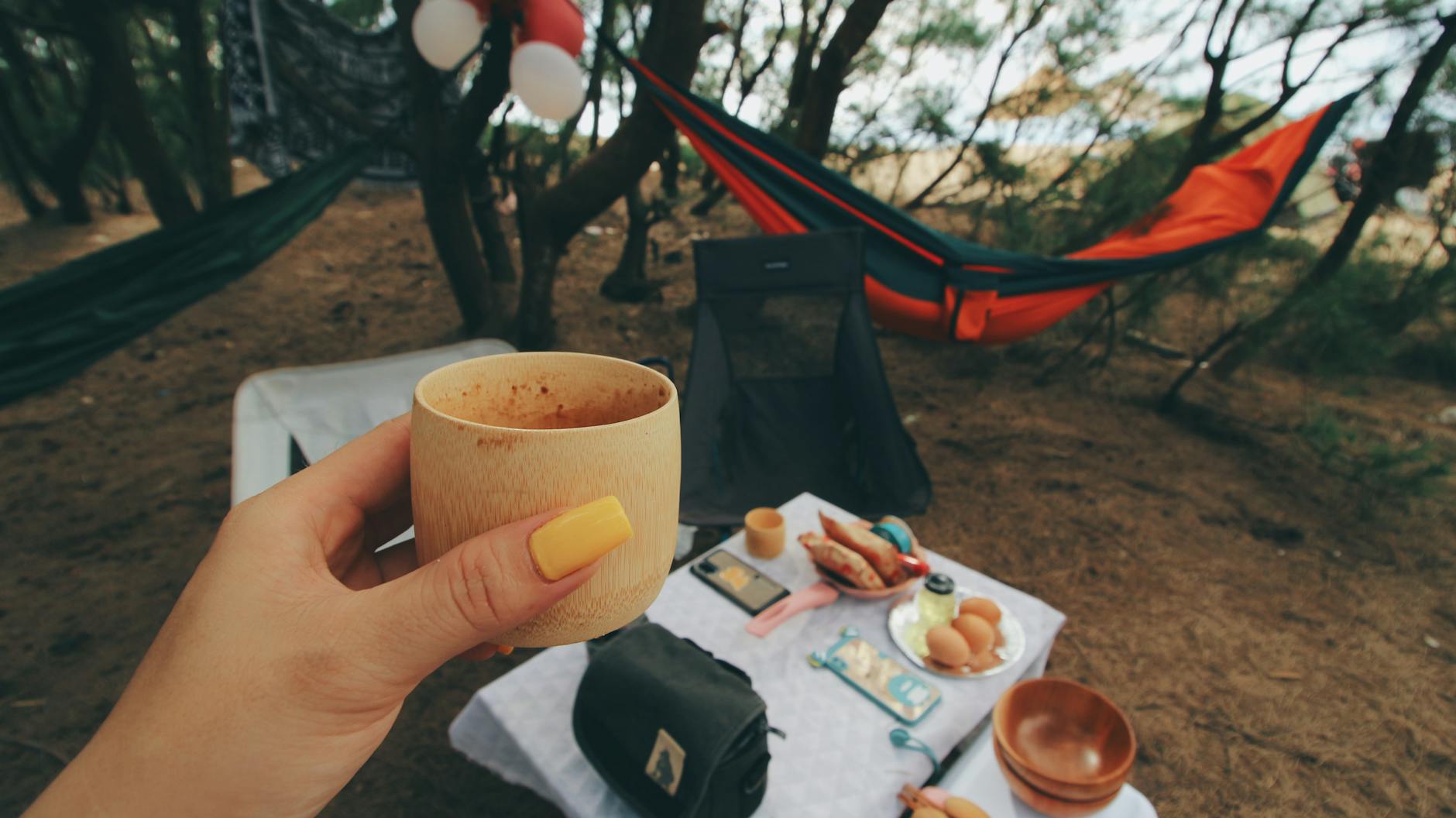 cozy camping setup with a hand holding a wooden cup hammocks and picnic items under trees