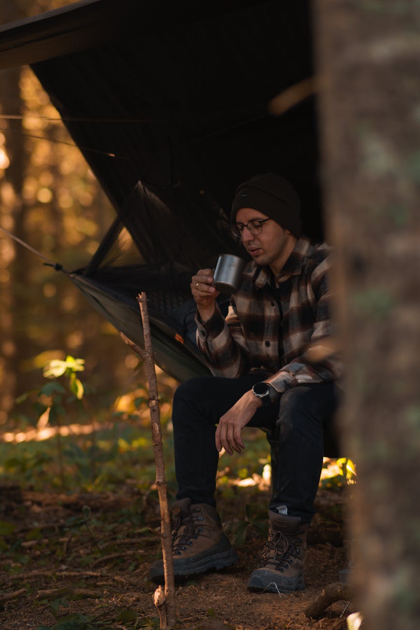 a man wearing a beanie and plaid shirt sipping coffee in a cozy forest camp setup