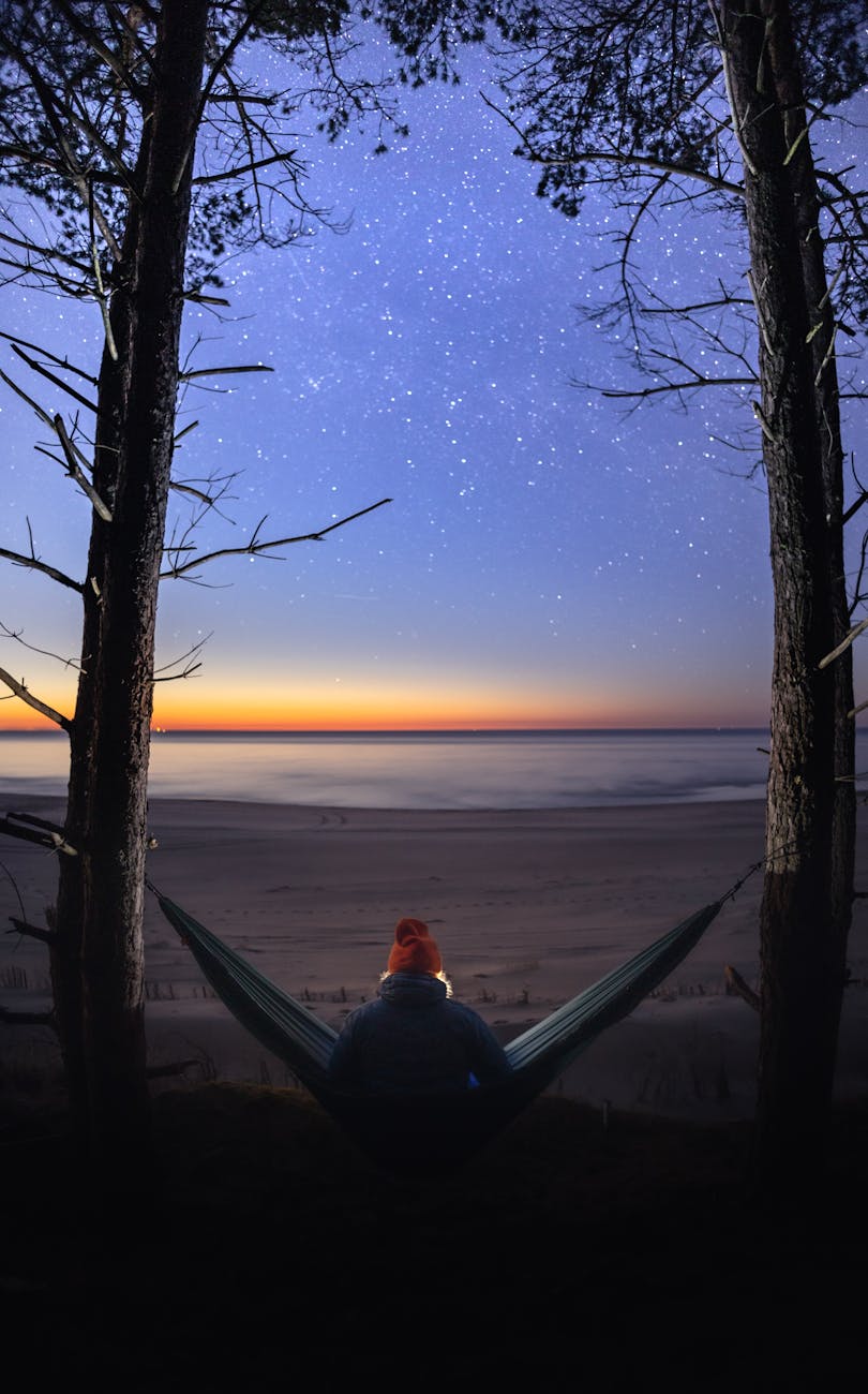 a person relaxing in a hammock framed by trees enjoying a stunning night sky over a serene beach