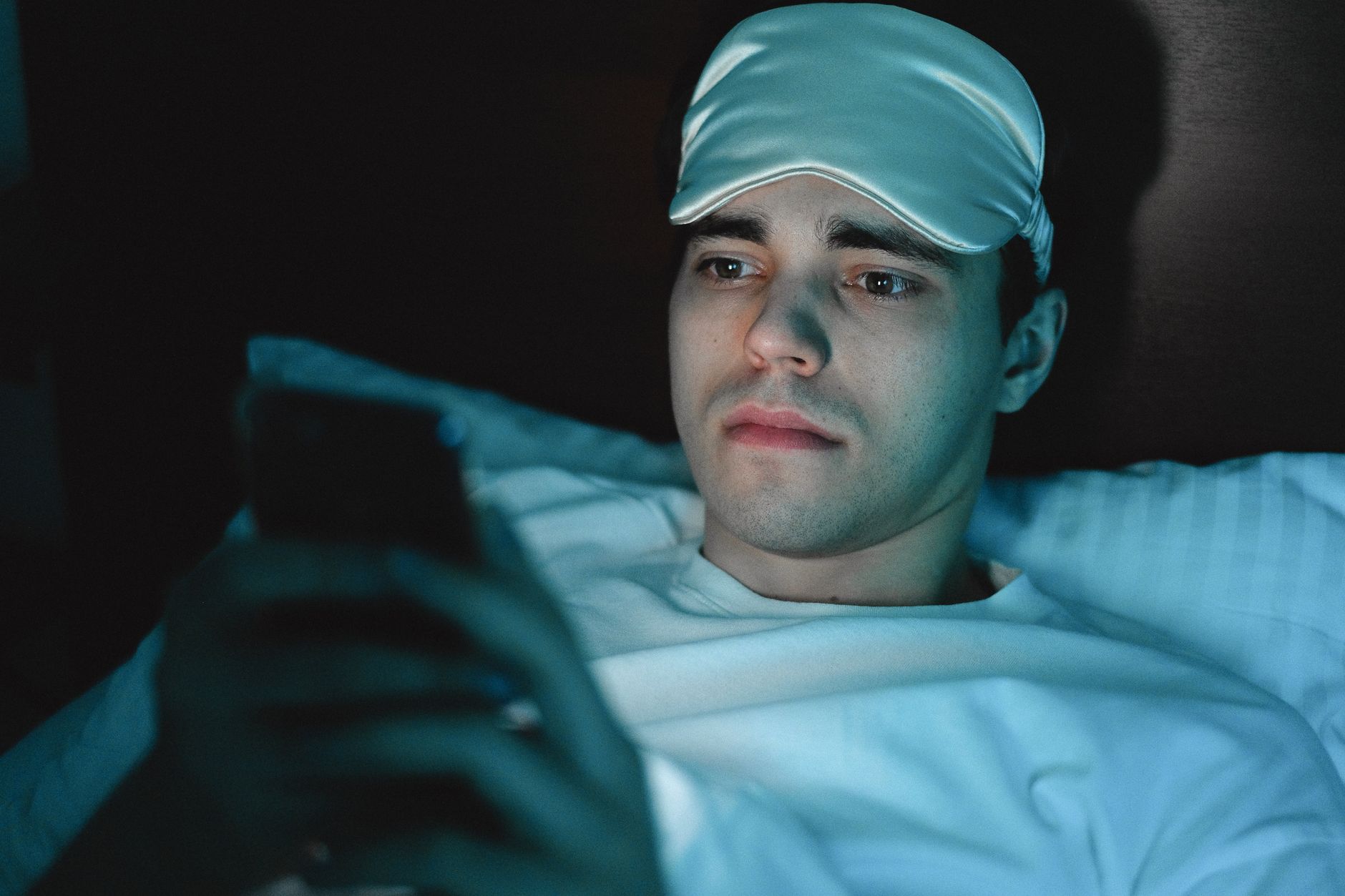 Un homme regarde son téléphone au lit, avec un masque de sommeil sur le front
