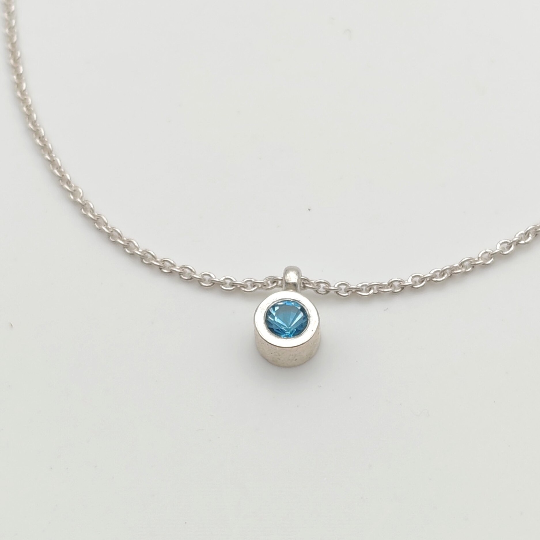 Vue rapprochée (gros plan) du pendentif en argent massif 925 orné dune topaze bleue fine à facettes. Serti clos artisanal, sur chaîne maille vénitienne. Photographie de détail sur fond blanc