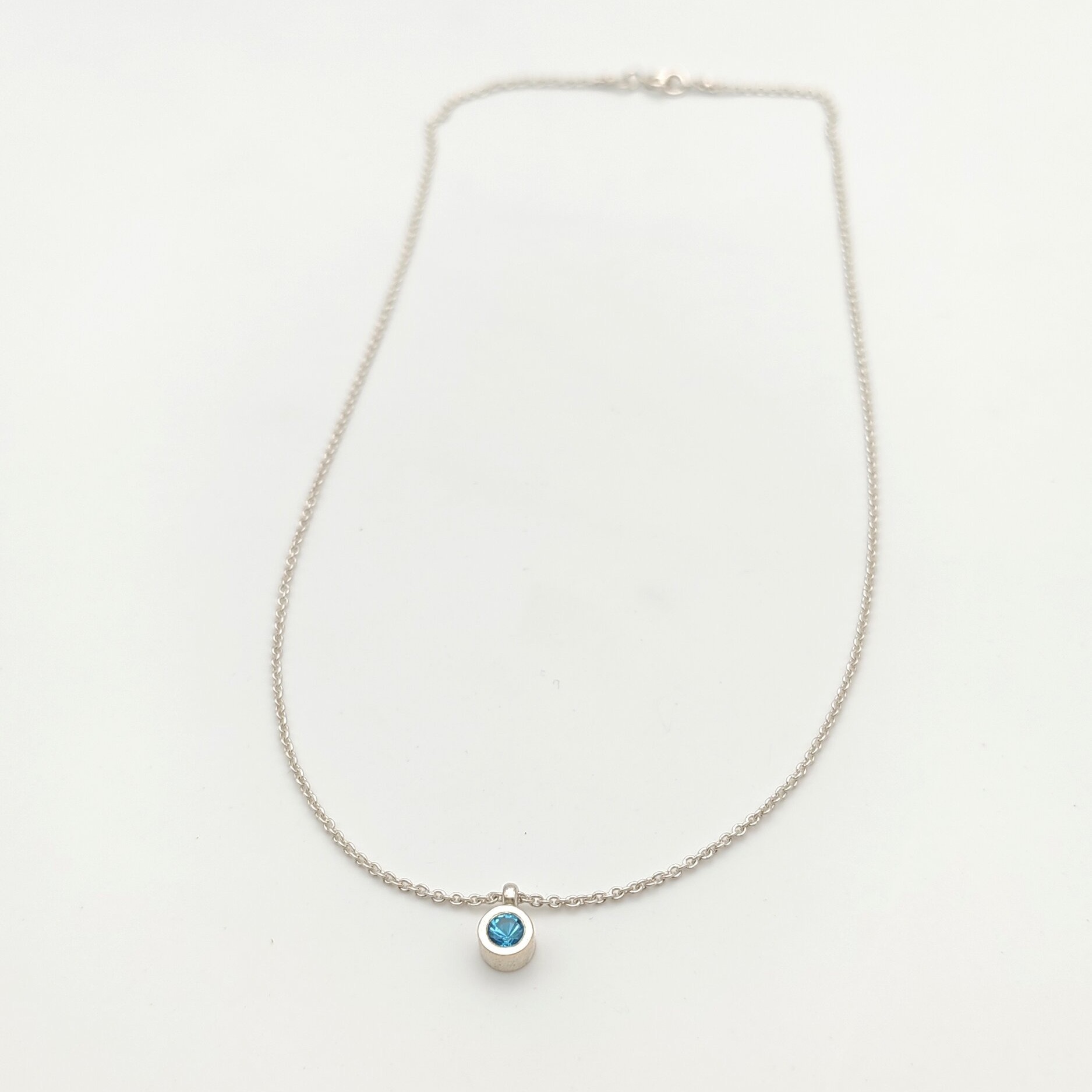 Photographie de produit professionnelle d'un collier pendentif en argent 925 avec topaze bleue ronde en serti clos sur fond blanc. Chaîne maille vénitienne de 41 cm. Élégance minimaliste