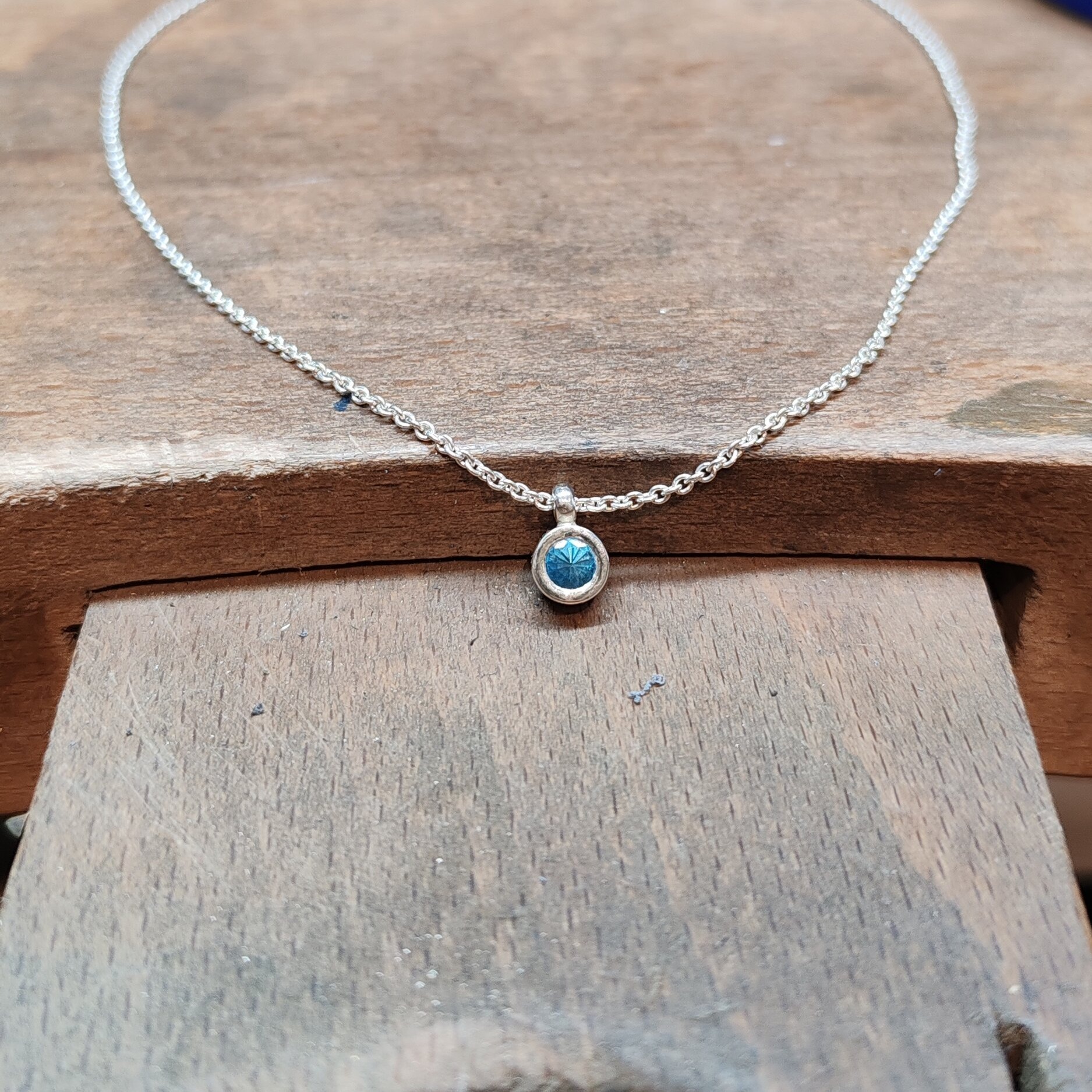 Collier pendentif en argent 925 avec topaze fine bleue et chaîne maille vénitienne posé sur létabli en bois dun bijoutier. Bijou artisanal moderne et raffiné en argent massif