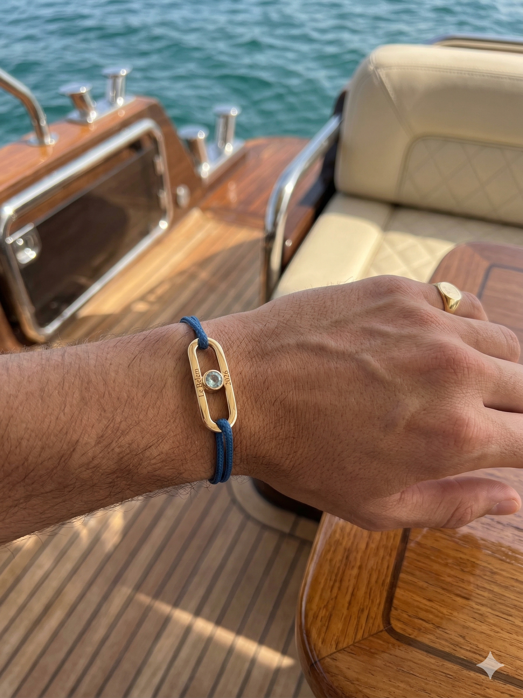 Bracelet Nautic Bijouterie JK en or 750 gravé, aigue-marine centrale et cordon bleu, porté par un homme sur le pont en teck d'un yacht.