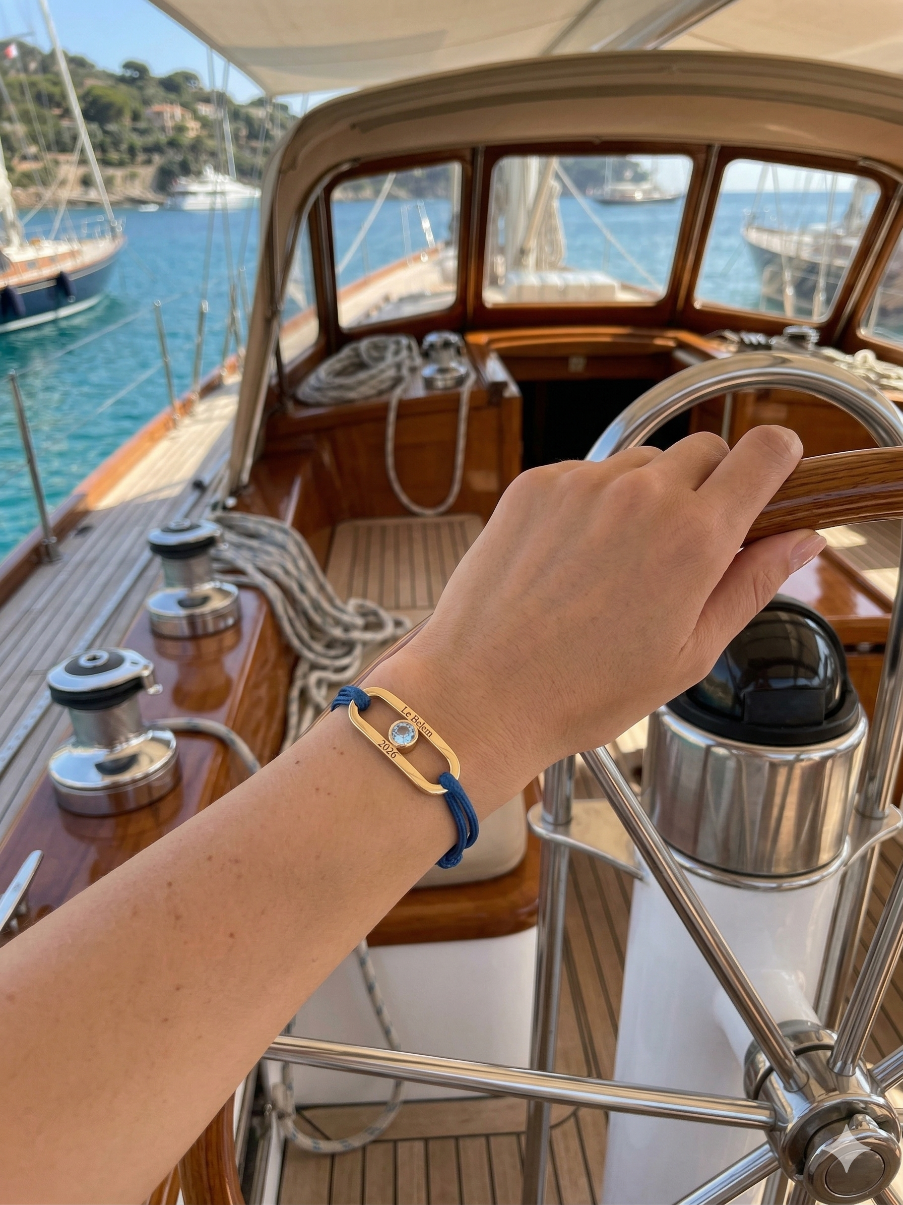 Bracelet Nautic Bijouterie JK en or 750 gravé, aigue-marine centrale et cordon bleu marine, porté par une femme sur un yacht près du volant.