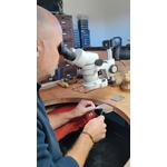 Lartisan bijoutier sertisseur de la Bijouterie JK en plein travail à son établi en bois. Il porte une veste bleu marine et utilise un binoculaire grossissant professionnel blanc pou