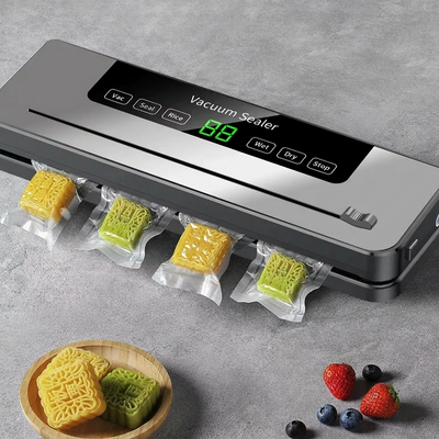 Machine d'emballage sous vide gris argenté sur surface gris béton. Gâteaux de lune jaunes et verts sous vide sortant de la machine. Écran LED vert affichant _88_. Panneau tactile _Vacuum Sealer_ avec boutons Vac,