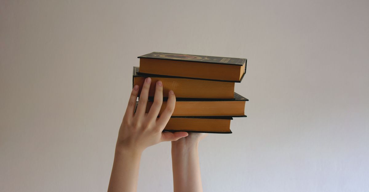 hands reaching upwards holding a stack of hardbound books against a plain background
