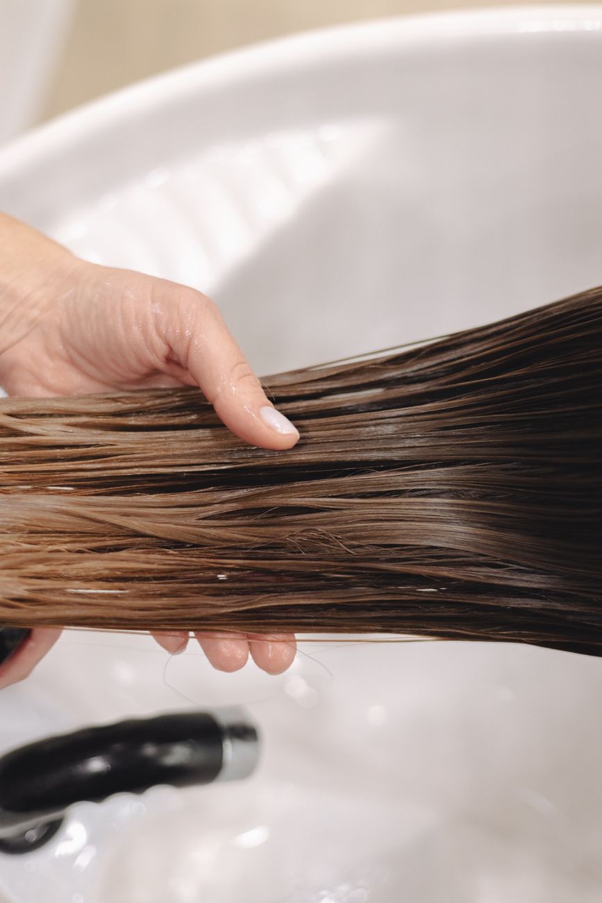 gros plan d une main tenant de longs cheveux bruns mouilles au dessus d un evier en ceramique blanc mettant en avant les soins capillaires