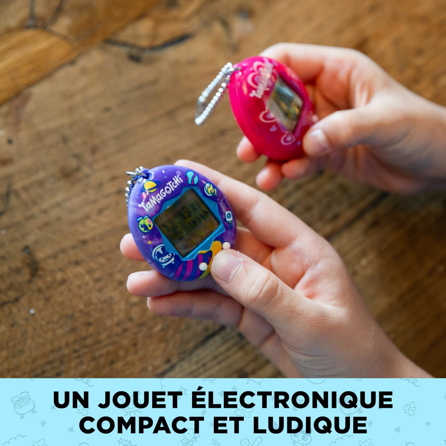 Deux mains tenant le Tama Universe et un Tamagotchi rose au-dessus dune table en bois