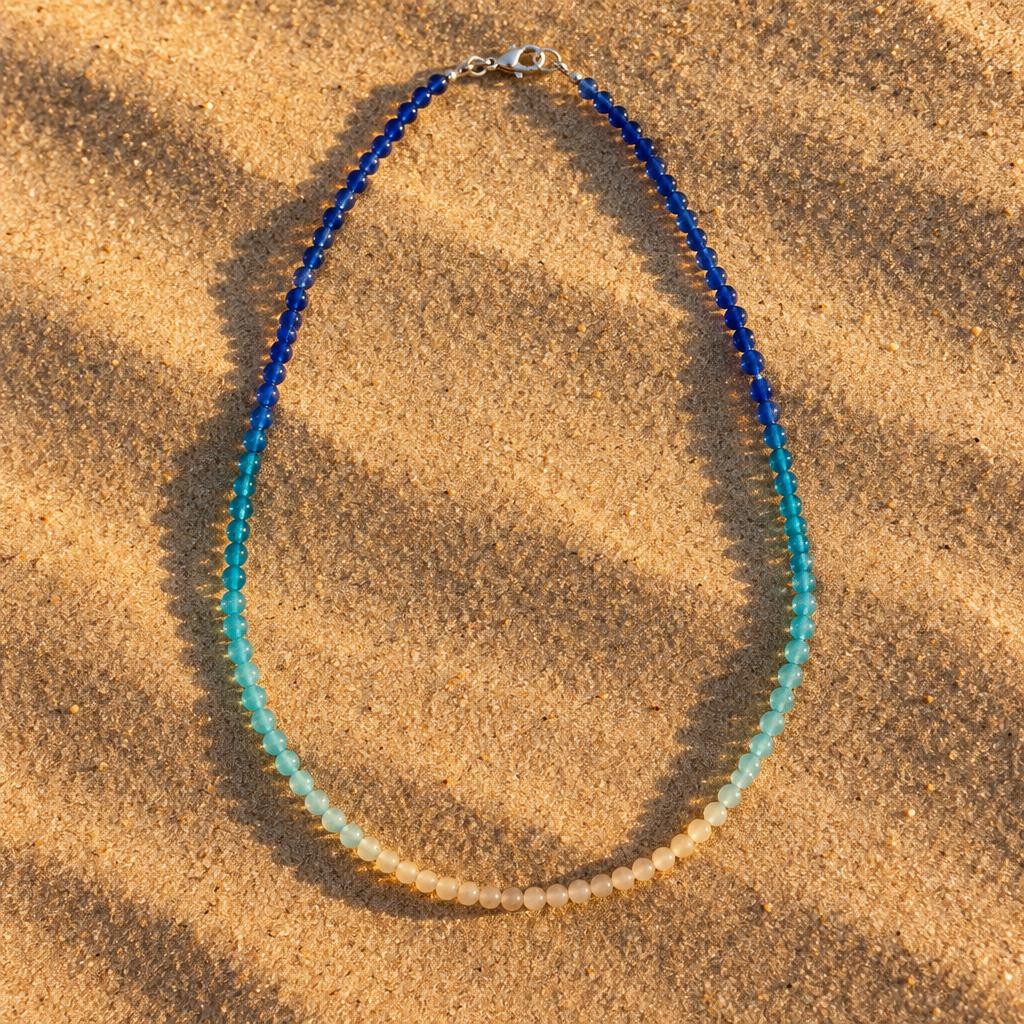 Collier en perles de verre en dégradé bleu océan et sable, posé sur le sable au coucher du soleil