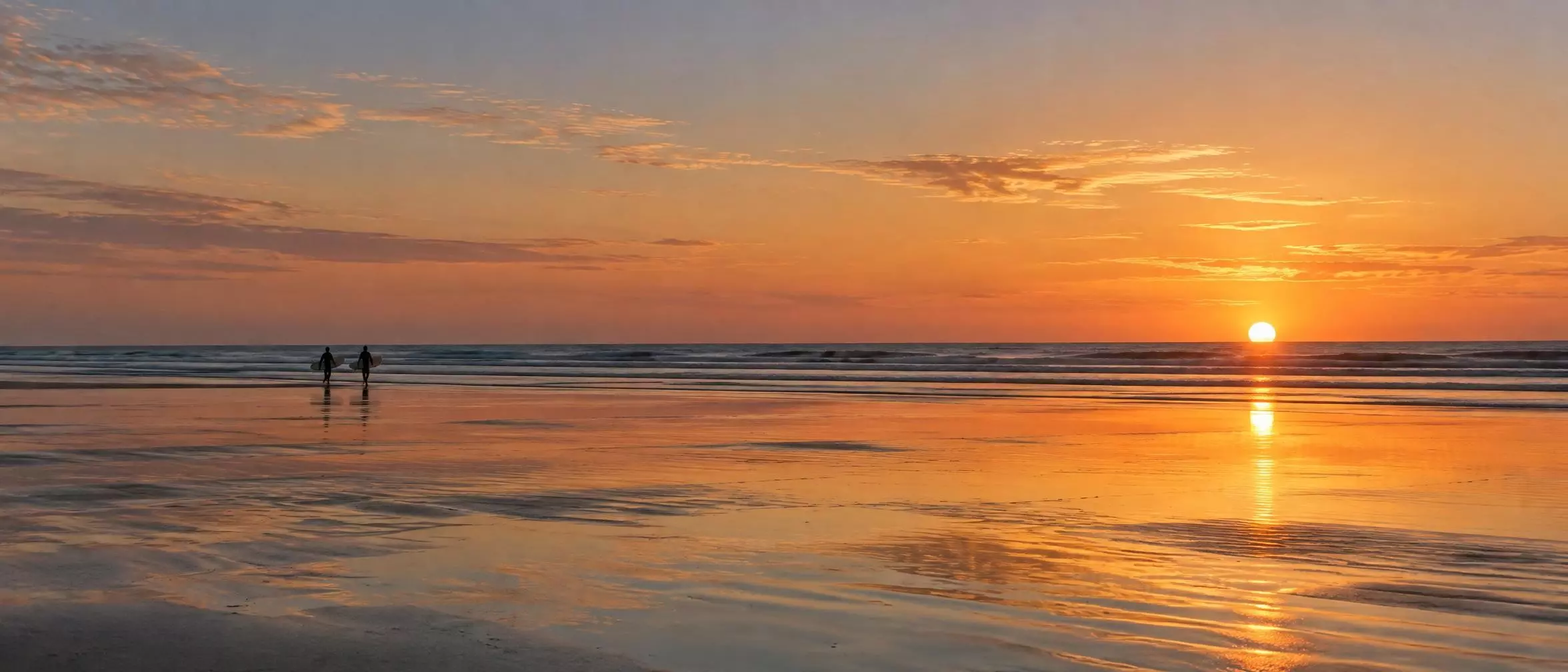 L’estran au coucher du soleil, sable mouillé miroir et deux surfeurs