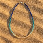 Collier en perles de verre en dégradé bleu océan et sable, posé sur le sable au coucher du soleil
