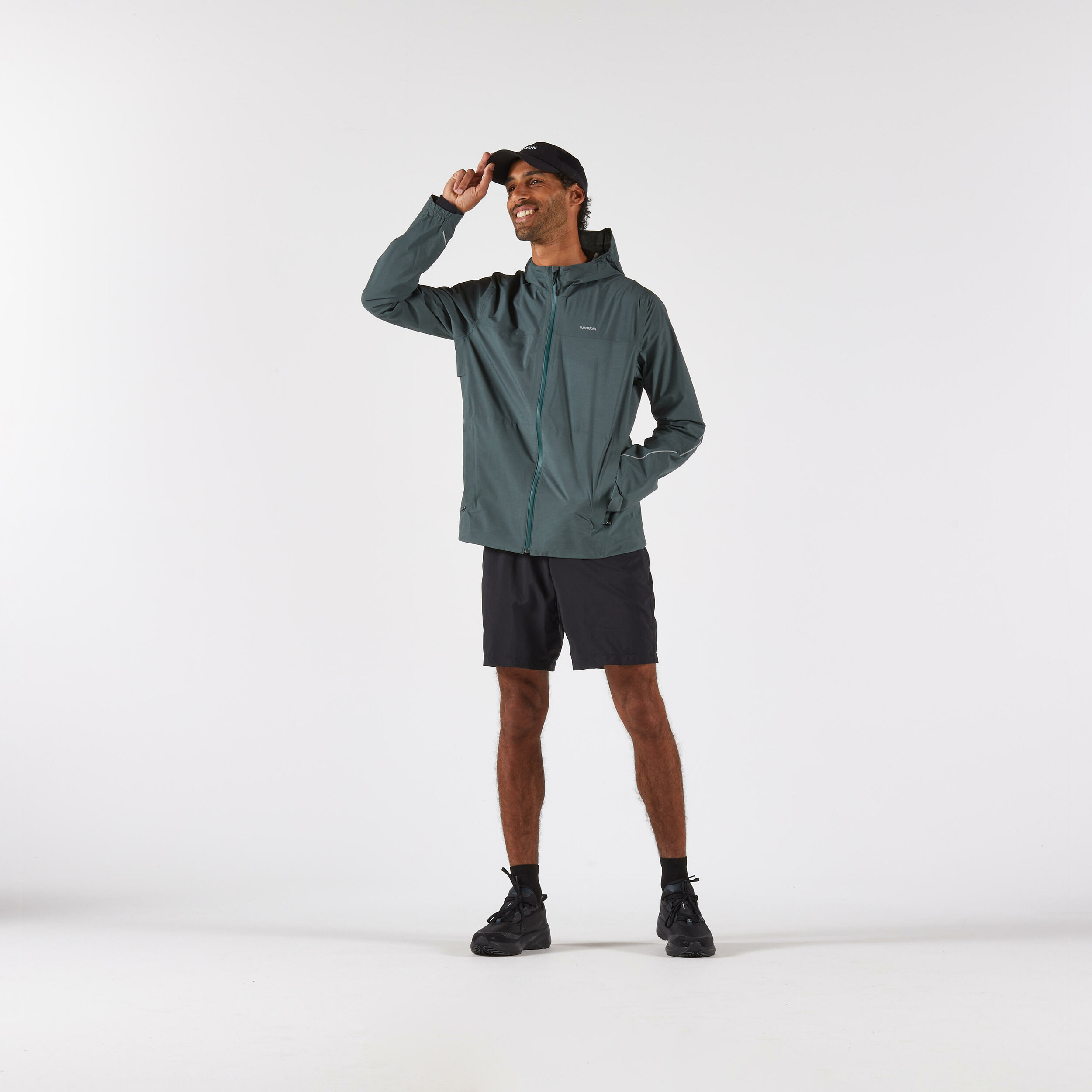 Runner in piedi con giacca Kiprun Run Flow verde militare, pantaloncini neri e cappellino