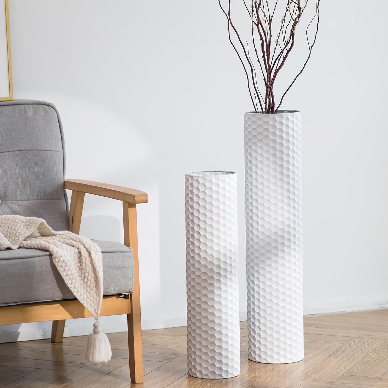 Zwei weiße Metallvasen mit Wabenmuster, 70 und 55 cm, mit dunklen Zweigen neben einem grauen Sessel
