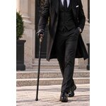 Homme élégant en costume noir marchant avec une canne épée à poignée tête de mort, devant un bâtiment de prestige.